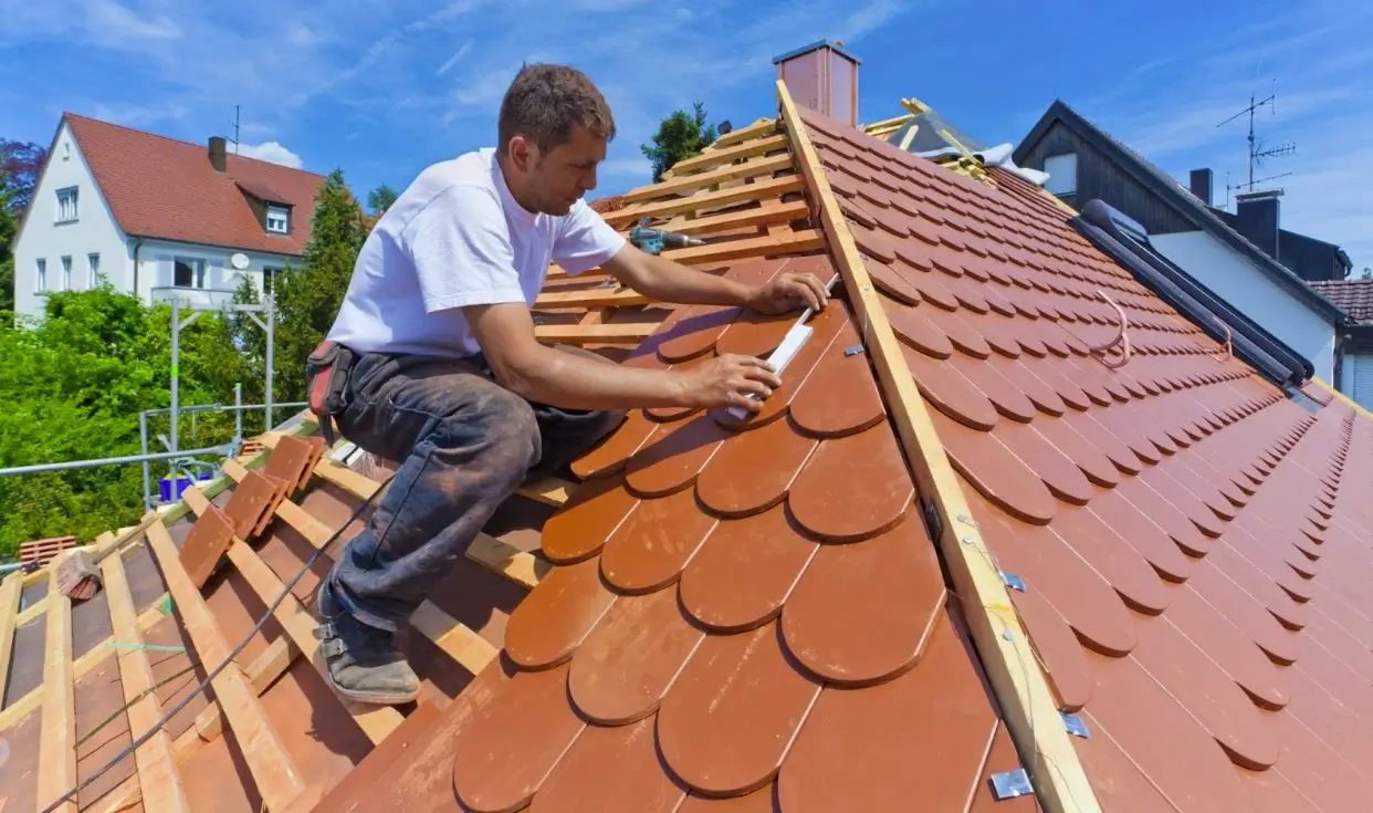 A roofer in a white shirt and jeans measures a tile atop a roof, with neighboring rooftops visible in the background, showcasing precision during restoration.