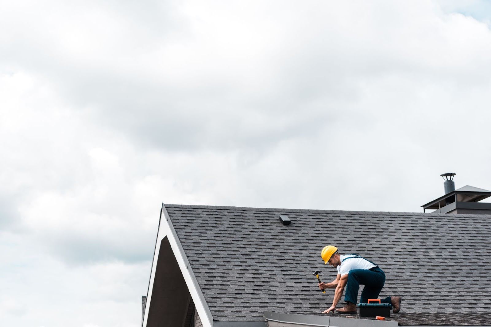 A roofer wearing a helmet fixes the roof shingles with a hammer.