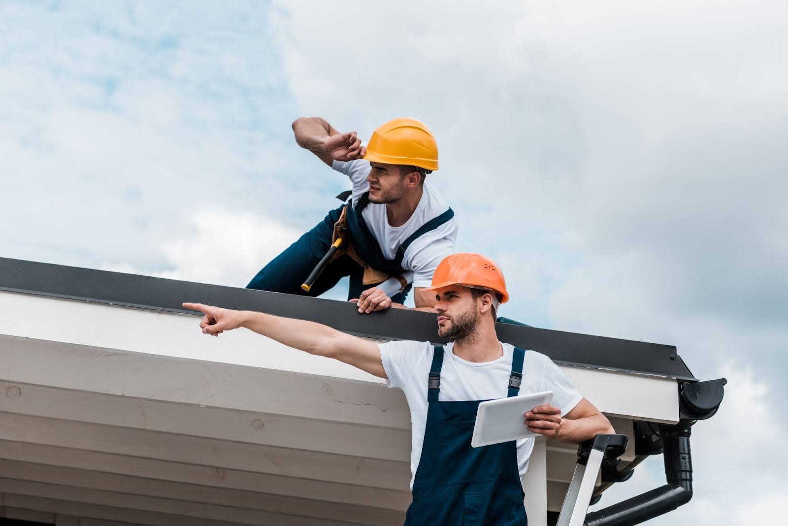 Bearded technician in safety gear reviews the roofing plans on a tablet and coordinates with a coworker.