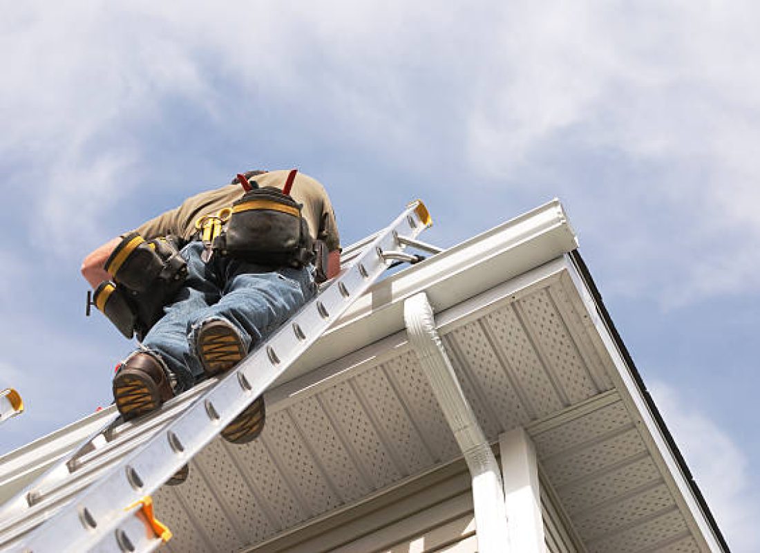 A handyman repairs his rain gutters.  He is up a ladder, photo taken from ground looking up, low angle view.  He wears a tool belt, sky and clouds, good copy space.
